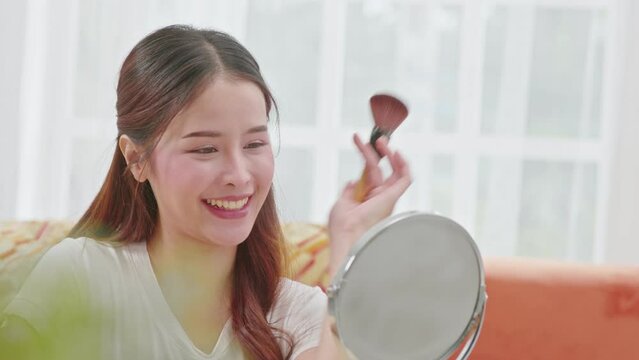 Happy Asian young woman smile and looking in mirror doing skin care applying make up brush on her cheek and beauty treatment at home,Wellness woman self care with beauty product at home