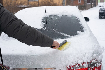 The hand of a man clearing snow from the rear window of a car with a brush.