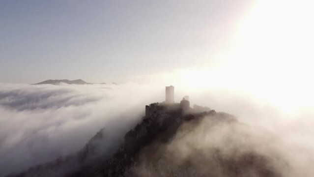 captivating drone zooms in on the majestic ruins of a castle atop a mountain. Mist weaves through ancient stones as the backlit sun creates a historic and mystical atmosphere in this compelling aerial