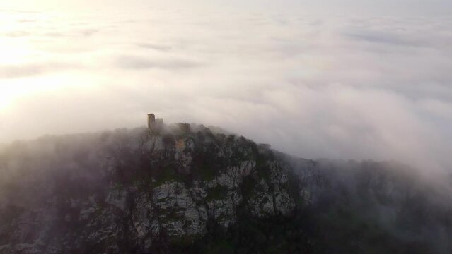 captivating drone zooms in on the majestic ruins of a castle atop a mountain. Mist weaves through ancient stones as the backlit sun creates a historic and mystical atmosphere in this compelling aerial