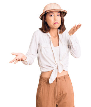 Young beautiful chinese girl wearing explorer hat clueless and confused expression with arms and hands raised. doubt concept.