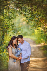 Fototapeta premium Beautiful, happy family on a walk in the park. Son, mom and dad. Mom holds the baby in her arms, dad hugs them tenderly. Dad looks at his newborn son. Happiness. Tenderness. Love. Newborn. Summer.