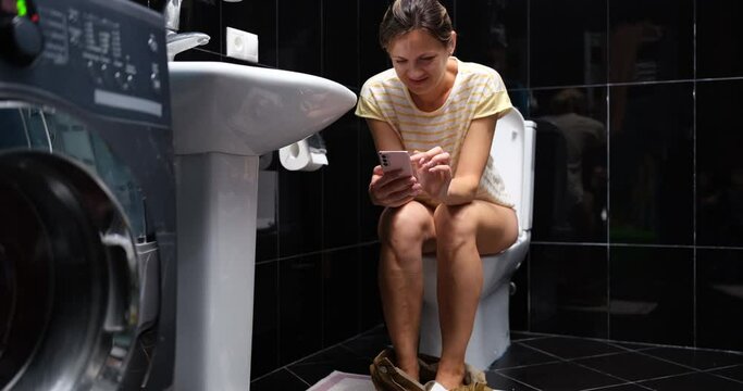 Woman using phone while sitting on toilet in the bathroom at home. Smartphone addiction and social media concept