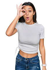 Fototapeta premium Young hispanic girl wearing casual white t shirt doing ok gesture shocked with surprised face, eye looking through fingers. unbelieving expression.