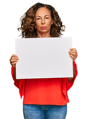 Middle age hispanic woman holding blank empty banner thinking attitude and sober expression looking...