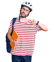 Young hispanic man wearing student backpack and bike helmet holding binder with angry face, negative sign showing dislike with thumbs down, rejection concept