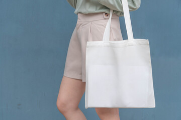 Blank white tote bag canvas fabric with handle mock up design. Close up of woman holding eco or reusable shopping bag against blue metal wall. No plastic bag and ecology concept.