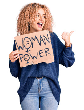 Young blonde woman with curly hair holding we need a change banner pointing thumb up to the side smiling happy with open mouth