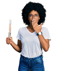 Young african american woman holding paintbrushes covering mouth with hand, shocked and afraid for...