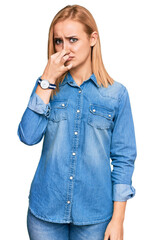 Beautiful caucasian woman wearing casual denim jacket smelling something stinky and disgusting, intolerable smell, holding breath with fingers on nose. bad smell
