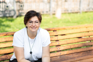 A beautiful middle-aged woman with glasses and short dark hair is sitting on a bench
