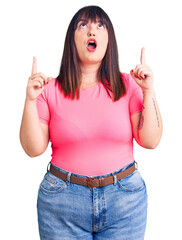 Young plus size woman wearing casual clothes amazed and surprised looking up and pointing with fingers and raised arms.