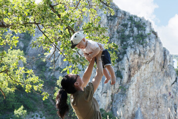 A beautiful woman and her little son are raised up in their arms against the backdrop of rocky mountains and trees. Summer portrait of mother and son on Mother's Day. Side view.