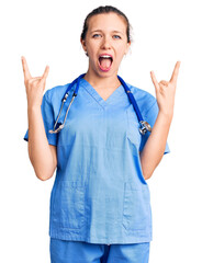 Young beautiful blonde woman wearing doctor uniform and stethoscope shouting with crazy expression doing rock symbol with hands up. music star. heavy concept.