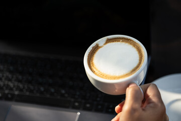 Business women hand holding white latte coffee cup and laptop on black table background.Hot coffee cup with laptop on desk when work from home.