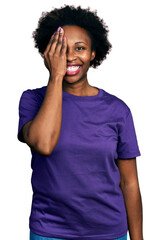 Fototapeta na wymiar African american woman with afro hair wearing casual purple t shirt covering one eye with hand, confident smile on face and surprise emotion.