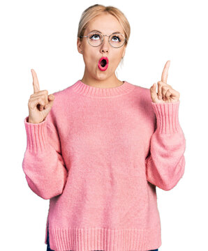 Young blonde woman wearing casual clothes and glasses amazed and surprised looking up and pointing with fingers and raised arms.