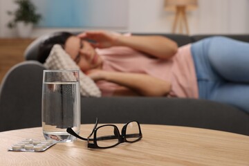 Sad woman suffering from headache indoors, focus on glass of water, pills and eyeglasses - obrazy, fototapety, plakaty