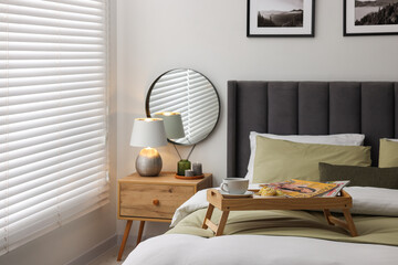 Wooden tray table with cup of drink and magazines on bed near window with horizontal blinds in room