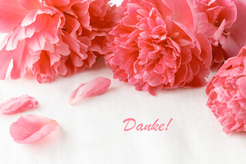 Danke (eng. Thank you) card. Pink coral peony flowers with falling petals on white linen table cloth.  Floral card with peony flowers and german word Danke.