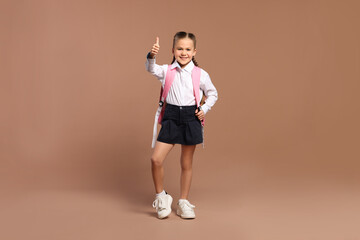 Happy schoolgirl with backpack showing thumb up gesture on brown background