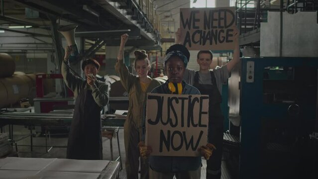 Medium shot of group of young multiethnic female factory workers with protest banners in hands shouting initiative slogans in mouthpiece while having strike during work shiftMedium shot of group of yo