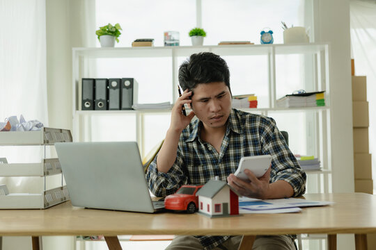 A young Asian man is stressed about bills, car payments, house payments, and holds his head in his hands as he looks at the bills. Confused about financial information or worried about spending money