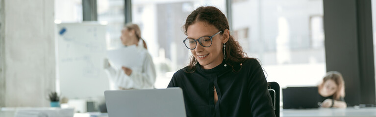 Smiling stylish businesswoman working on laptop sitting in modern office on colleagues background