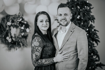 black and white photo of a smiling couple embracing, dressed in formal attire with a Christmas tree in the background.