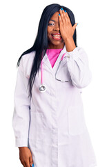 Young african american woman wearing doctor stethoscope covering one eye with hand, confident smile on face and surprise emotion.