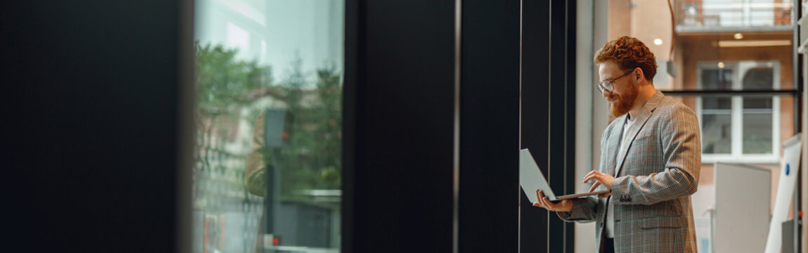 Focused male sales manager in eyeglasses working on laptop standing in office during working day