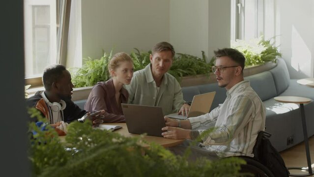 Young man in wheelchair showing business project on laptop and discussing it with team of multi-ethnic colleagues during informal office meetingYoung man in wheelchair showing business project on lapt