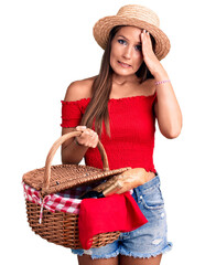 Young beautiful hispanic woman wearing summer hat and holding picnic wicker basket with bread stressed and frustrated with hand on head, surprised and angry face