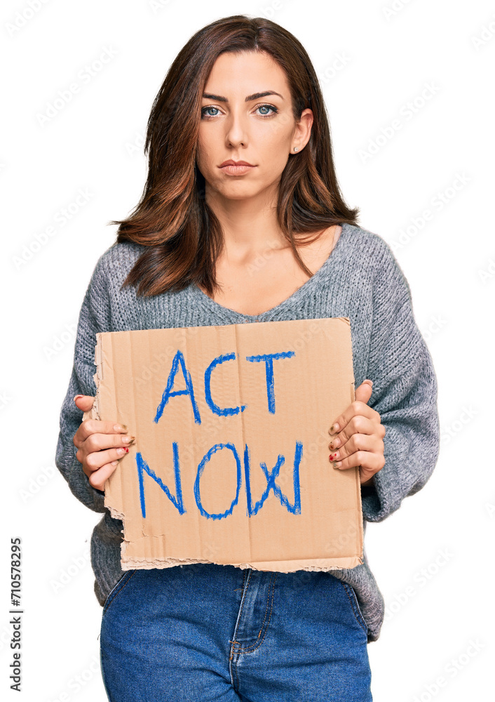 Wall mural Young brunette woman holding act now banner thinking attitude and sober expression looking self confident