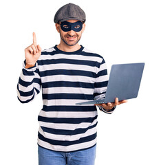 Young handsome man wearing burglar mask using laptop surprised with an idea or question pointing finger with happy face, number one