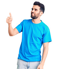 Naklejka premium Young handsome man with beard wearing casual t-shirt looking proud, smiling doing thumbs up gesture to the side
