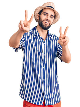 Young handsome man with beard wearing summer hat and striped shirt smiling looking to the camera showing fingers doing victory sign. number two.