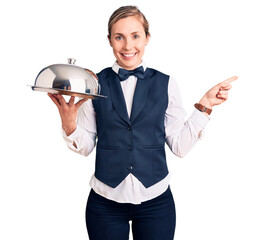 Young beautiful blonde woman wearing waitress uniform holding tray smiling happy pointing with hand...