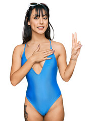 Young hispanic woman wearing swimsuit and sunglasses smiling swearing with hand on chest and...