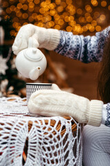 Hands in mittens pour tea into a cup against the background of the house