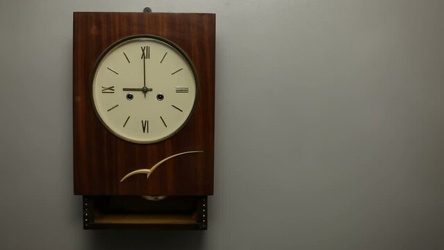 A large retro style wall clock with Roman numerals and a pendulum on a gray wall strikes exactly 9 o'clock. The video is looped and endless with sound. Old wall clock.