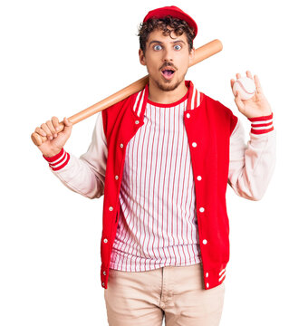 Young handsome man with curly hair playing baseball holding bat and ball scared and amazed with open mouth for surprise, disbelief face