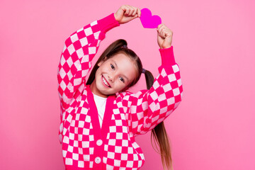 Photo of toothy beaming cheerful schoolgirl with tails dressed knit cardigan hold paper heart over...
