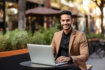 Happy young Latin business man using laptop computer sitting outdoor. Smiling Hispanic guy student or professional looking away in city cafe elearning, hybrid working