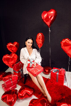 brunette woman in a white shirt on the background of balloons of hearts unpacks gifts. The concept of Birthday and Valentine's Day.