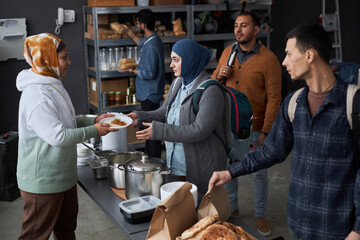 High angle view at group of Middle Eastern people receiving hot meals at soup kitchen or refugee support center with volunteer helping - obrazy, fototapety, plakaty