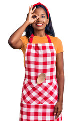 Obraz na płótnie Canvas Young indian girl wearing professional baker apron doing ok gesture with hand smiling, eye looking through fingers with happy face.