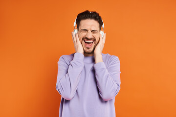 excited bearded man in purple sweater and wireless headphones listening music on orange background