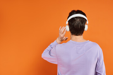 back view of man in purple sweater and wireless headphones listening music on orange background