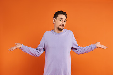 confused bearded man in purple sweater showing shrug gesture with his hands on orange background - obrazy, fototapety, plakaty